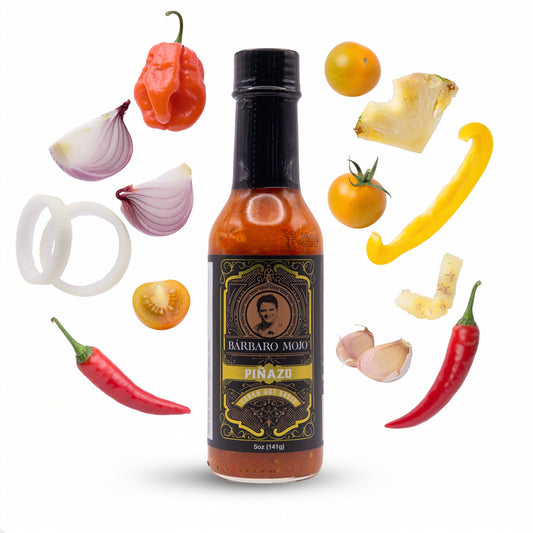 Barbaro Mojo Piñazo bottle surrounded by pineapple, yellow tomatoes, habanero, Fresno peppers, yellow bell pepper, onion and garlic