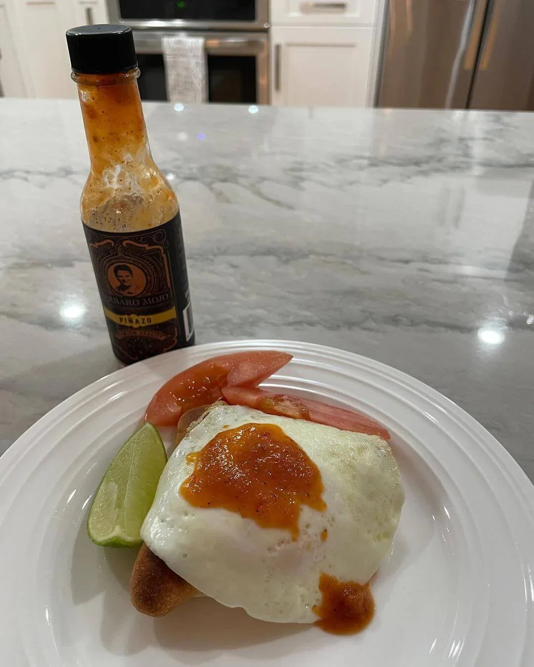 Plate with a fried egg topped with Barbaro Mojo hot sauce, tomato and lime, bottle in the background