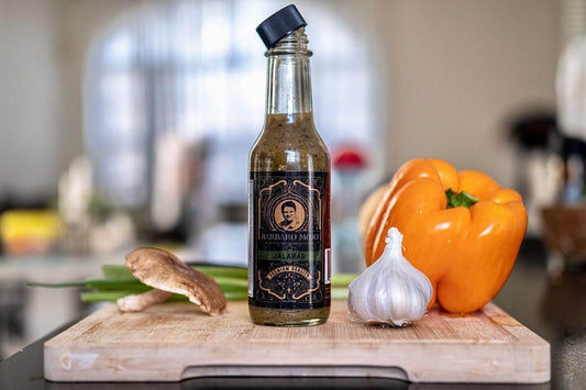 Bottle of Barbaro Mojo Cuban hot sauce on a cutting board with an orange bell pepper, garlic and a mushroom