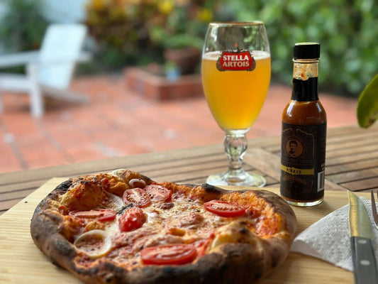 Wood-fired pizza with tomato and mozzarella on a cutting board next to a glass of beer and a bottle of Barbaro Mojo hot sauce, outdoors