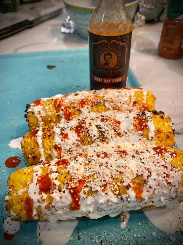 Roasted corn on the cob topped with crema, cheese and Barbaro Mojo Best Day Ever hot sauce, bottle in the background